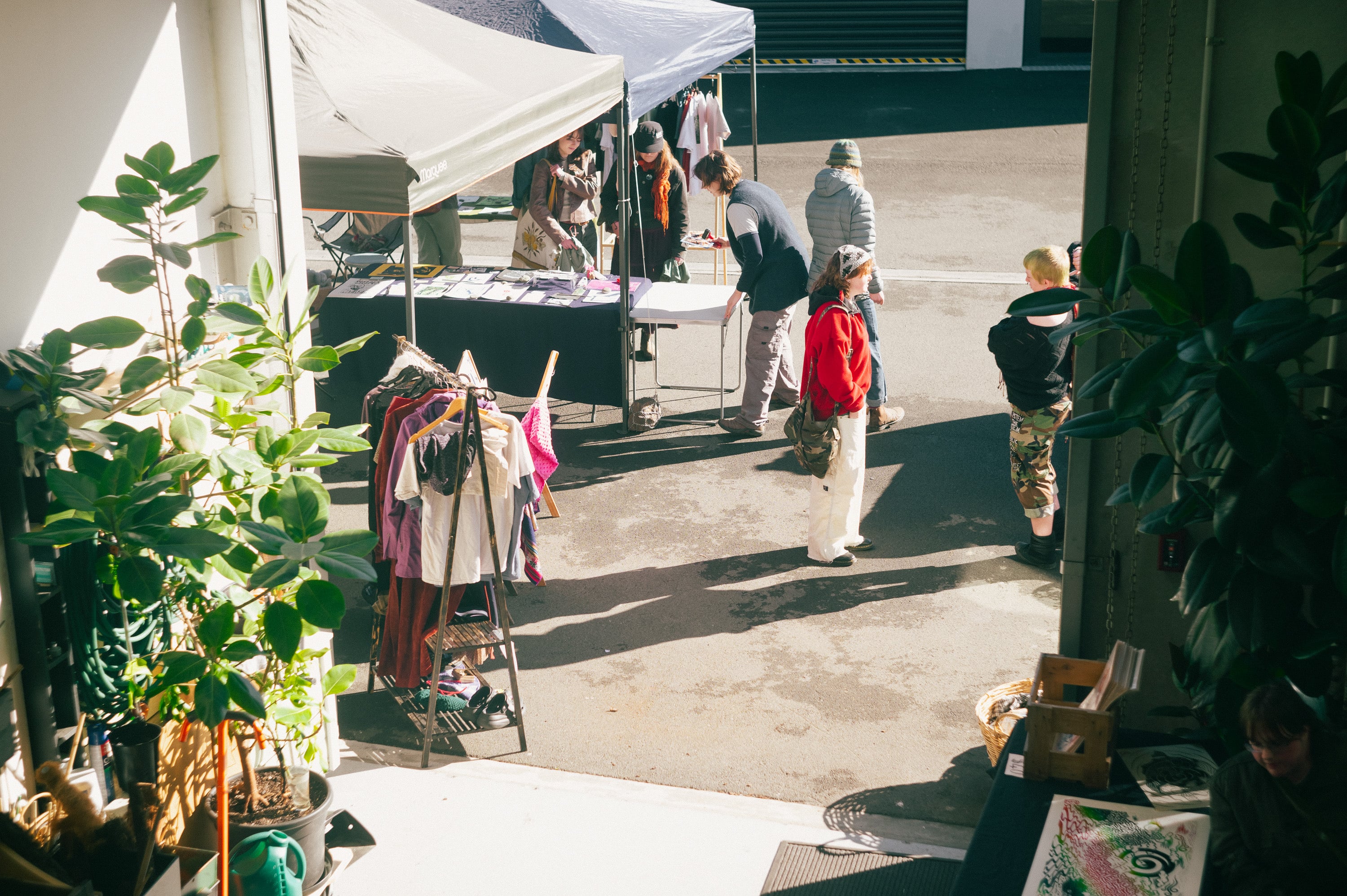 Sunlit market stalls with plants and racks of clothing.