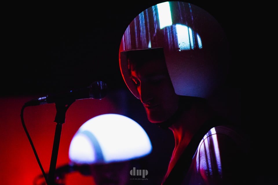Performer in a mirrored visor beside a glowing lamp, under red light.