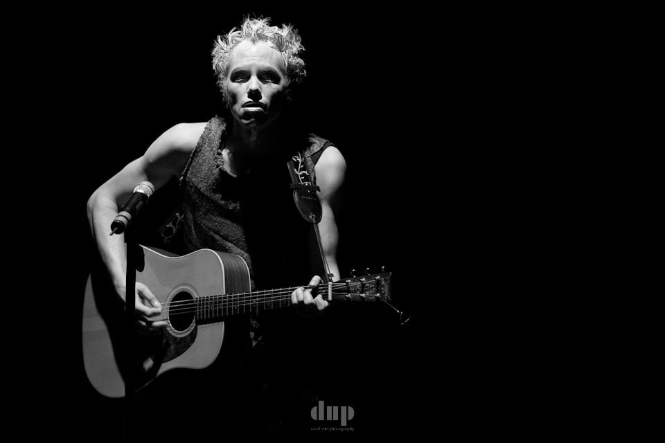 Singer-songwriter with an acoustic guitar under a single spotlight, black and white.
