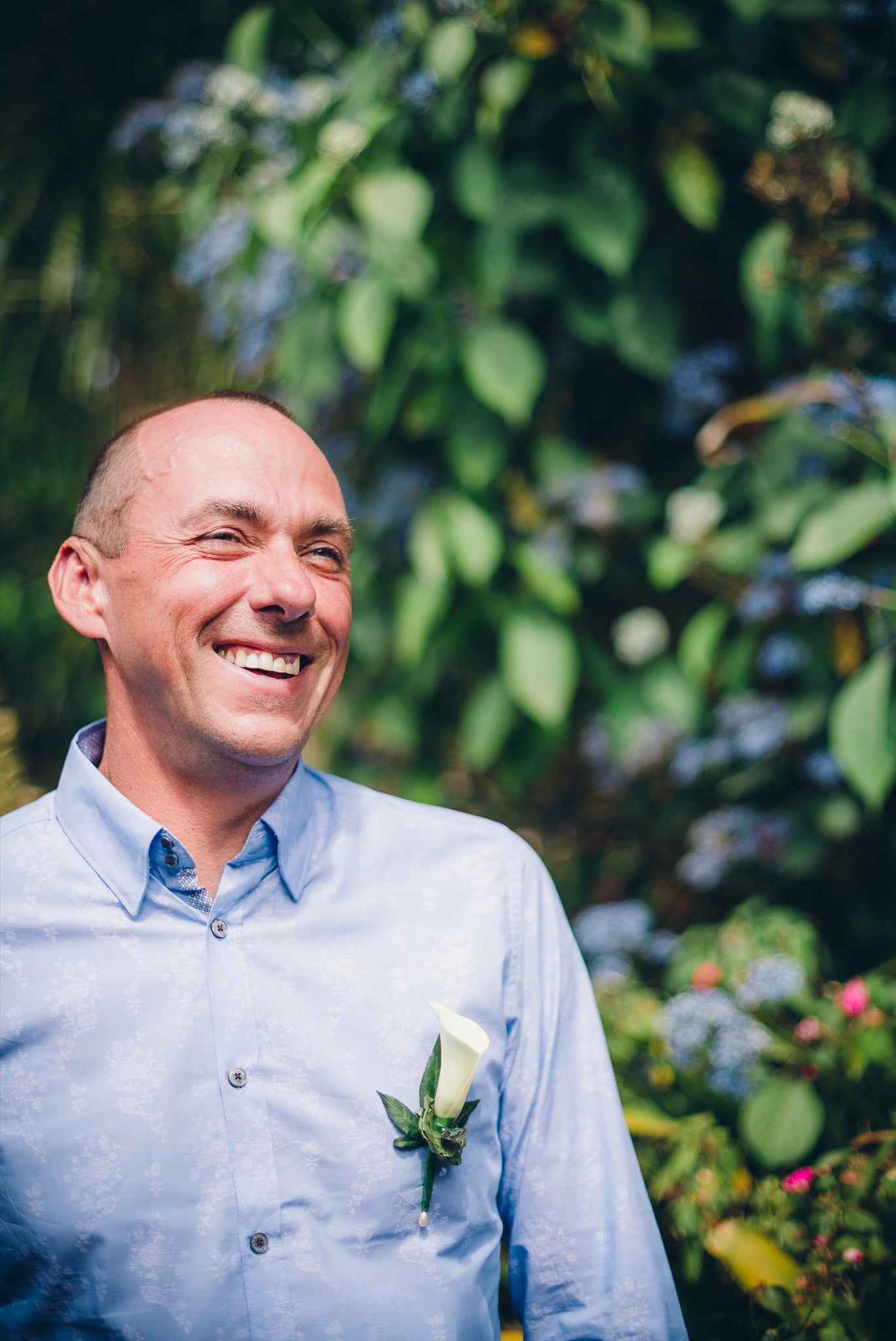 Man laughing in a garden, flower pinned to his shirt.