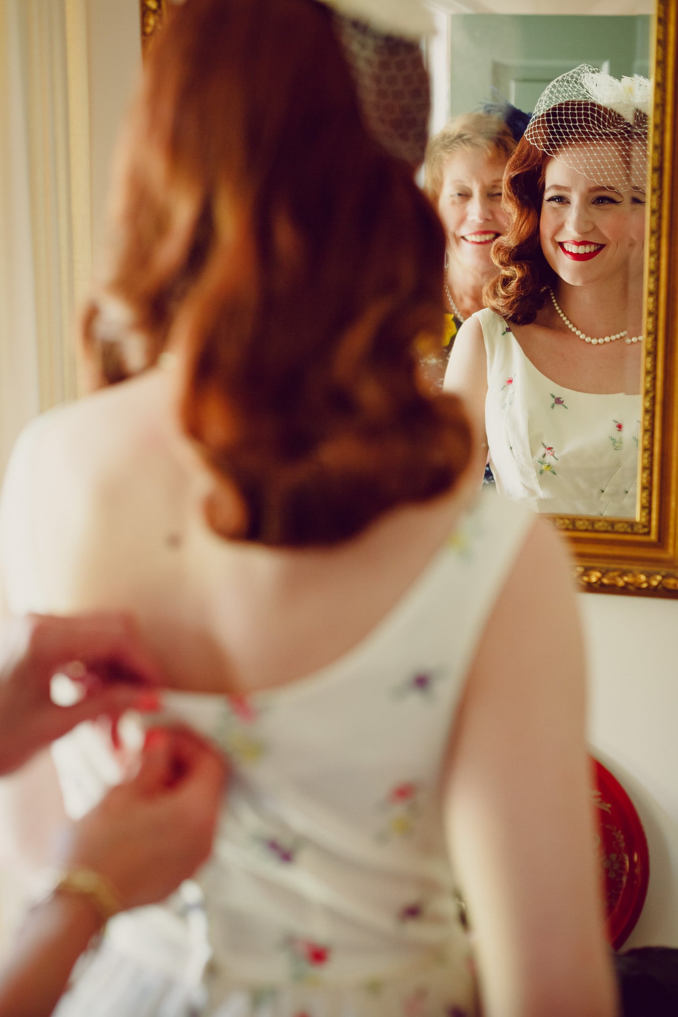 Bride being buttoned into a vintage dress, reflected in a mirror.