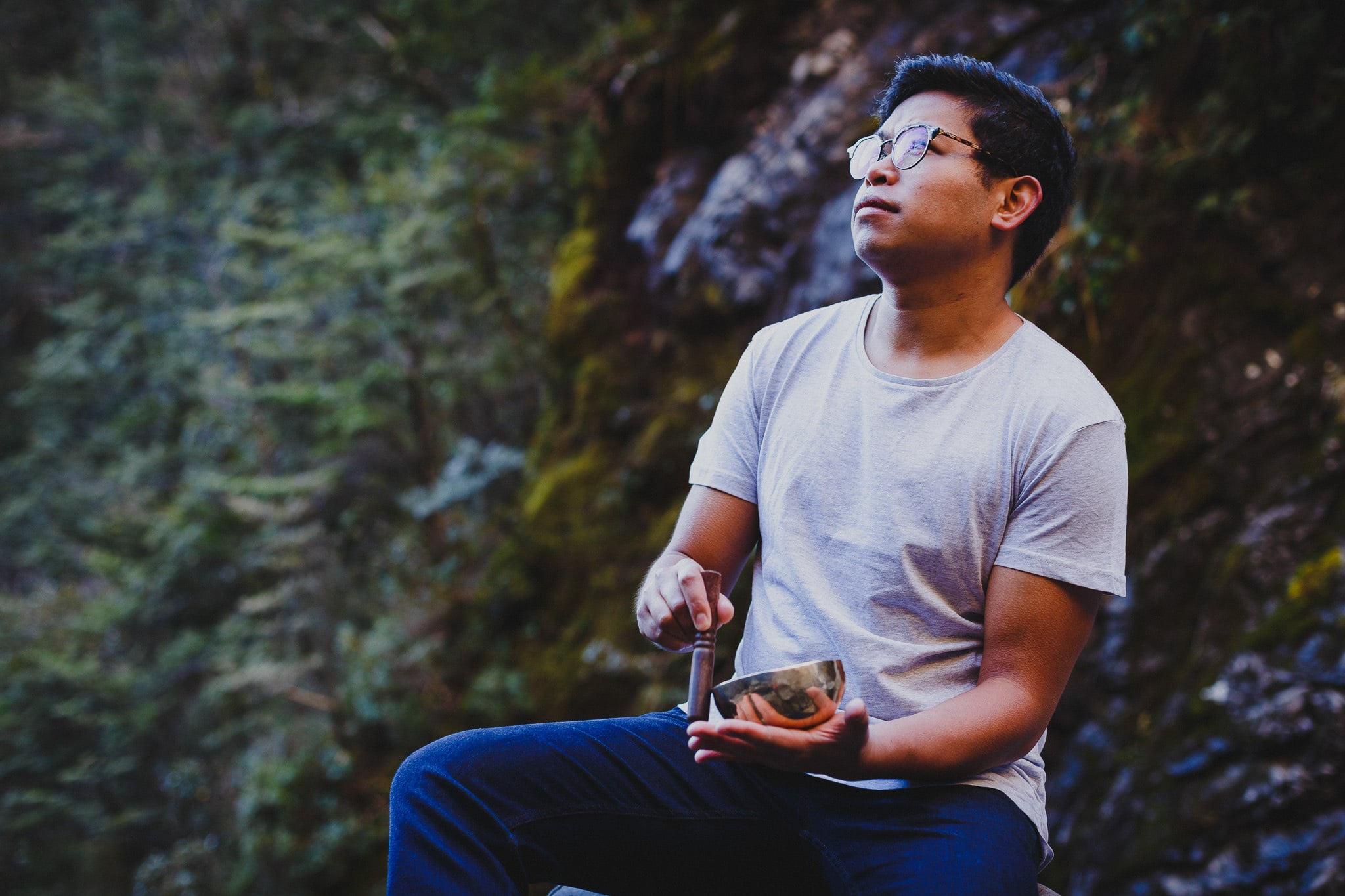 Man with glasses playing a singing bowl against a mossy rock face.