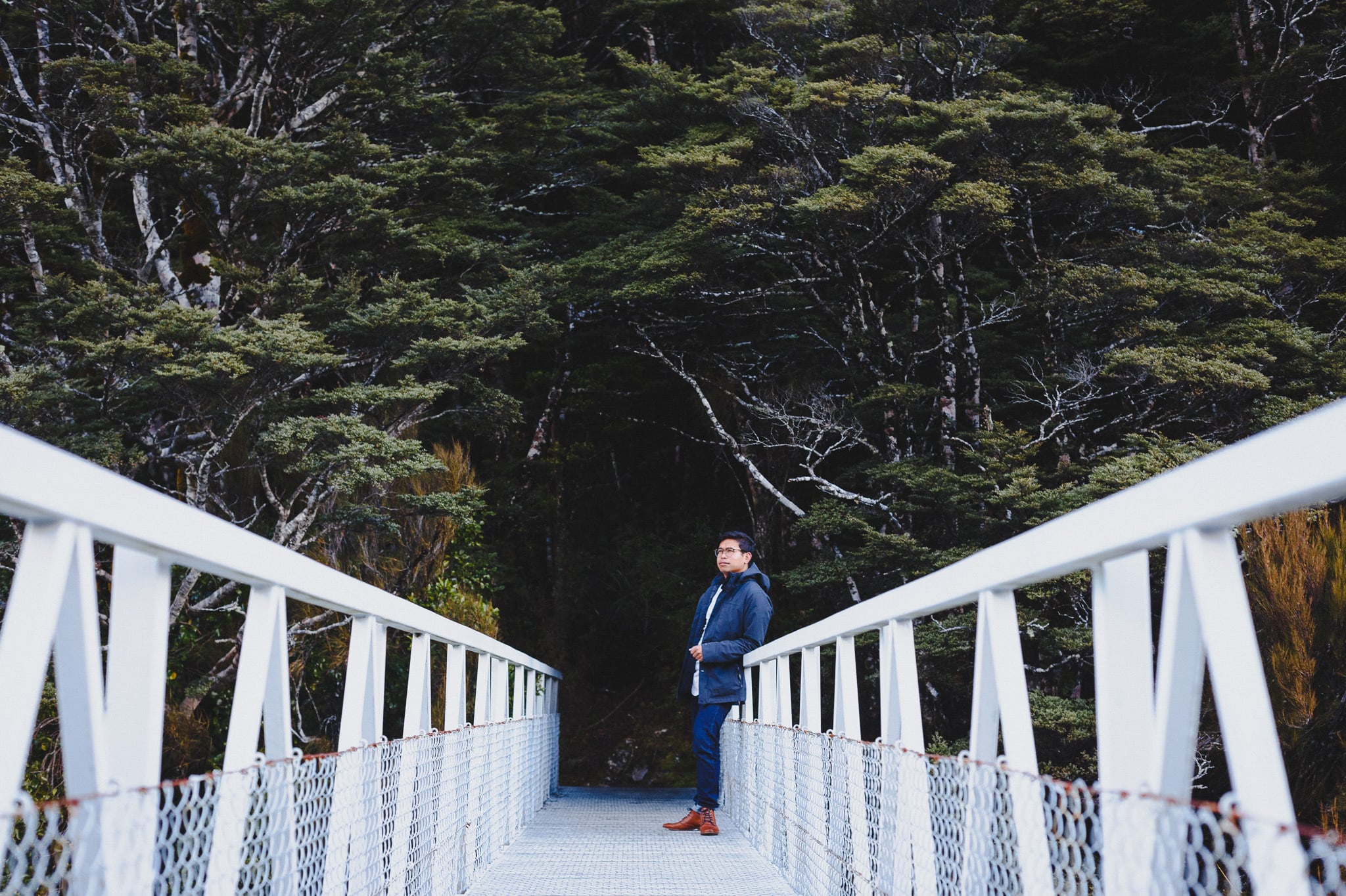 Man on a long white footbridge leading into dark forest.