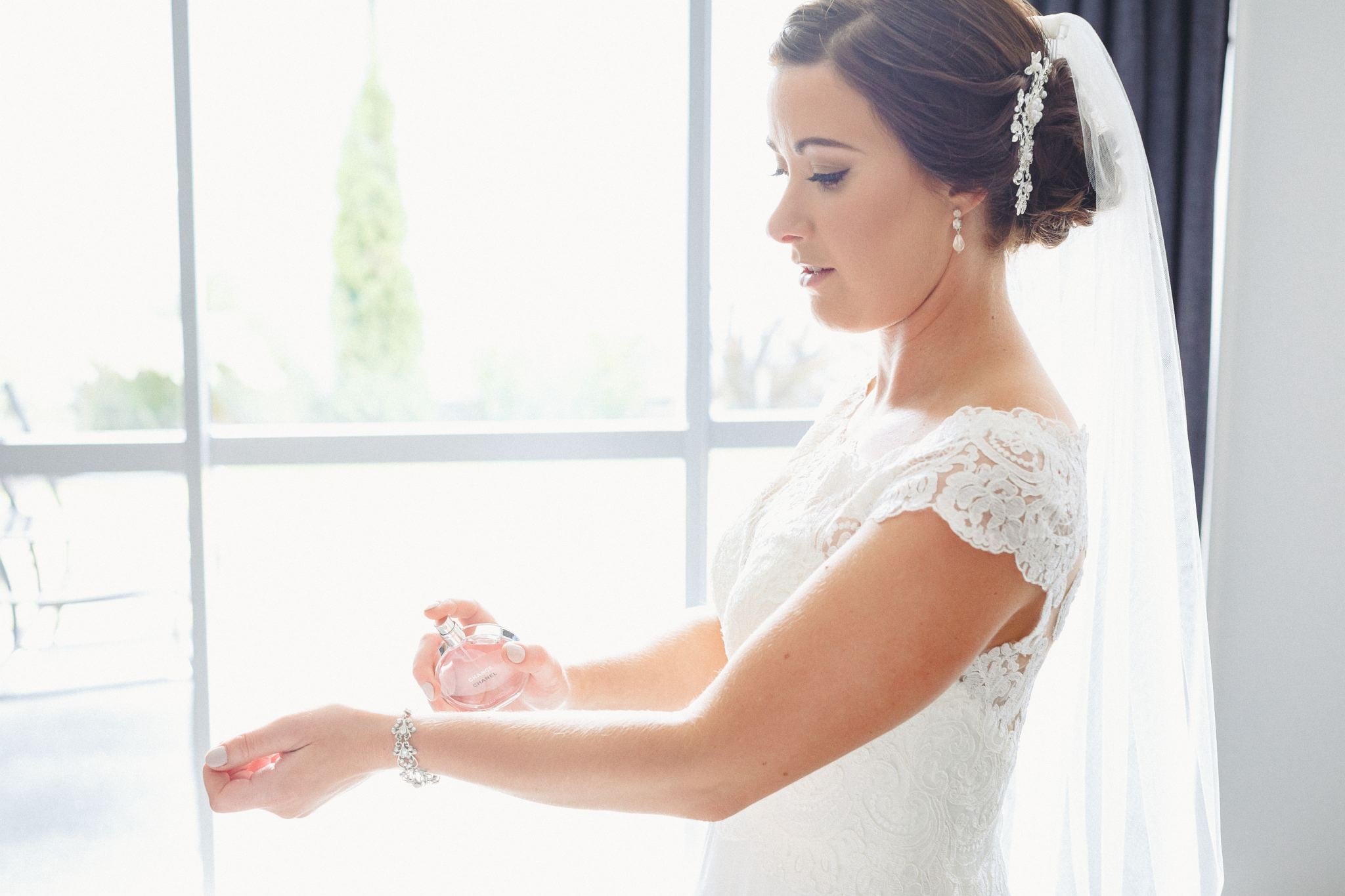 Bride putting on perfume by a bright window.