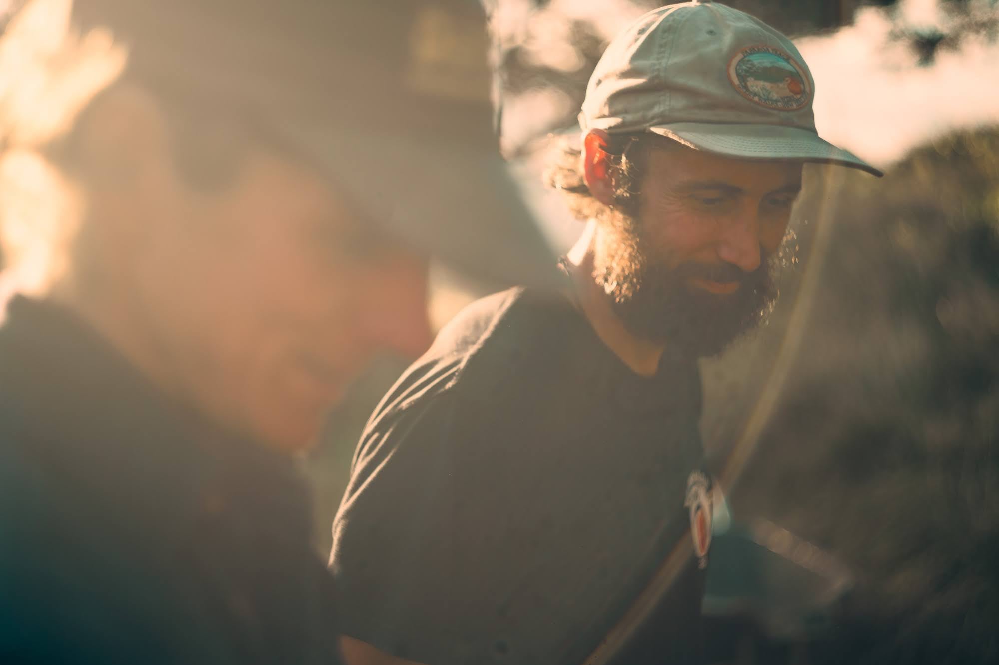 Bearded man in a cap looking down, softly backlit.