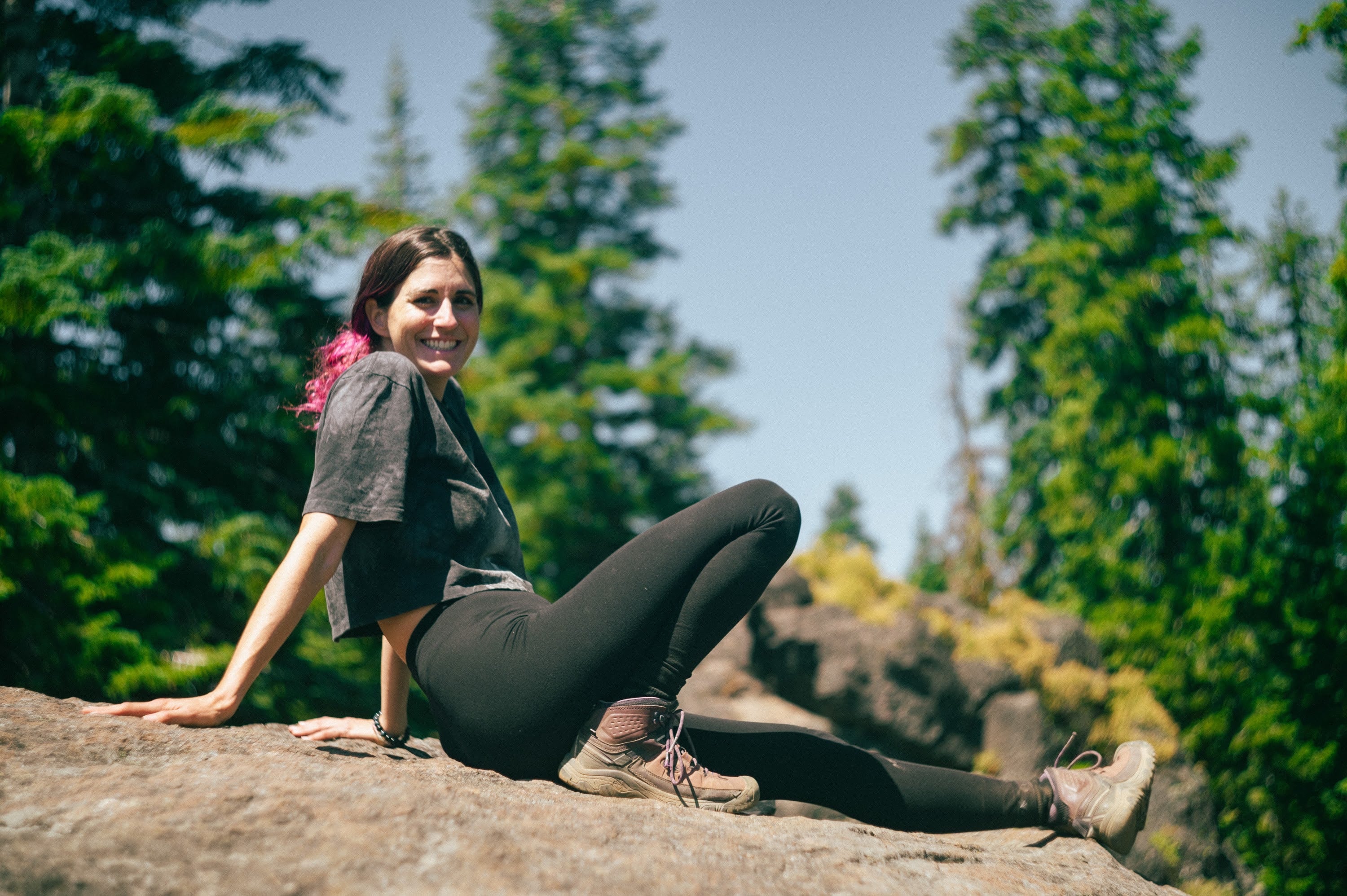 Woman with pink-streaked hair smiling from a sunny rock, pine trees behind.