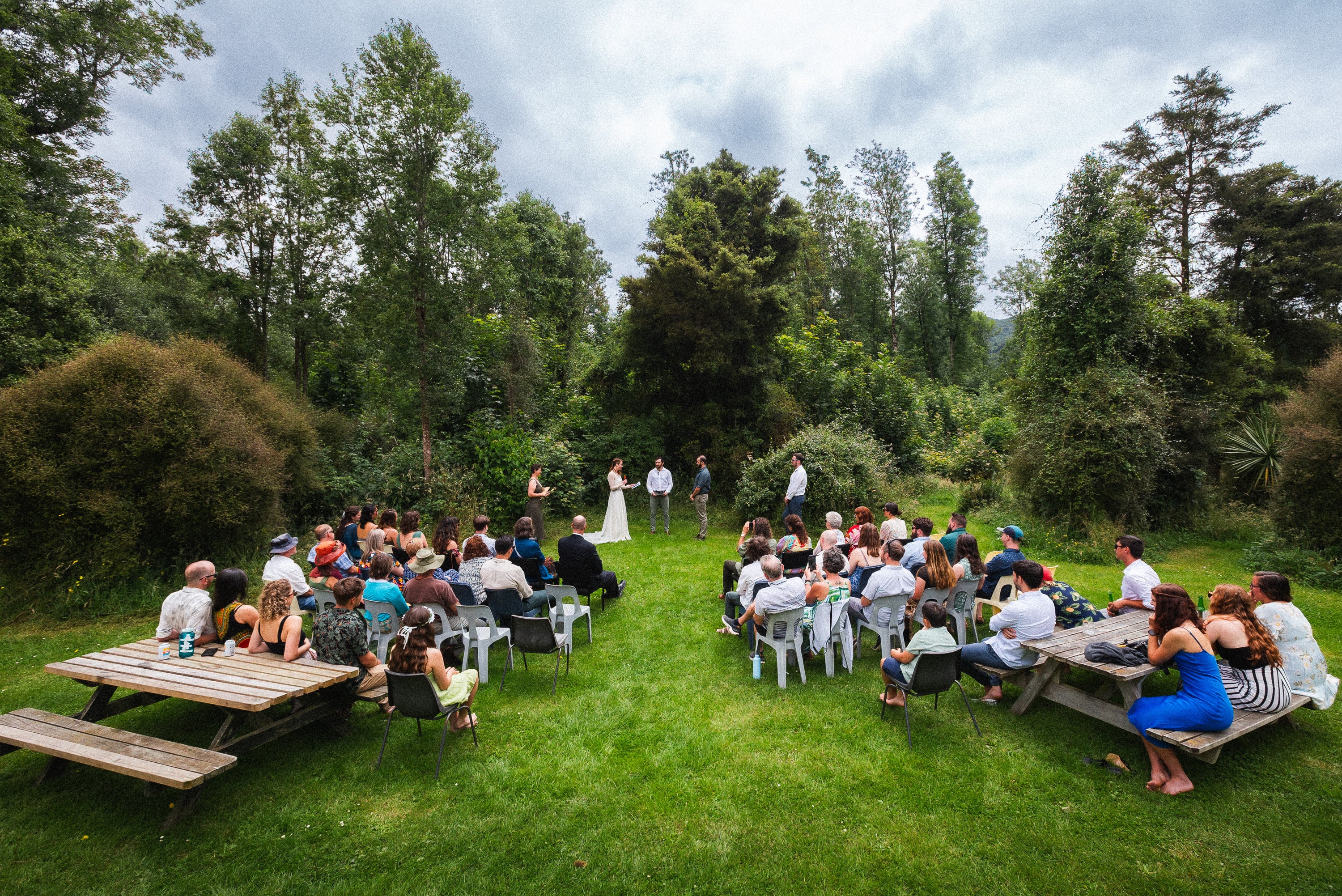 Outdoor ceremony in a forest clearing, guests at picnic tables.