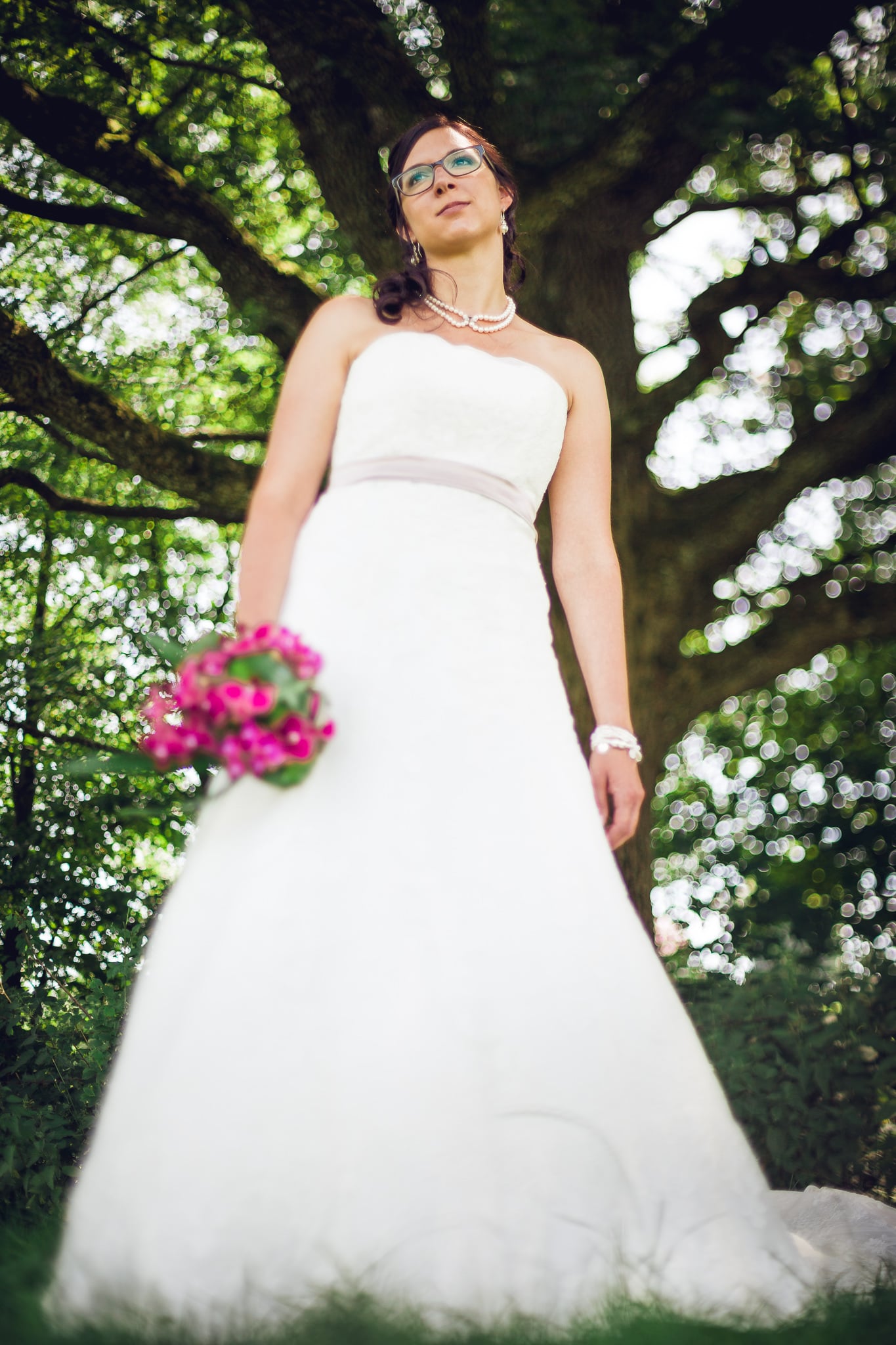 Bride standing under a tree with a pink bouquet.